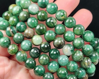 May include: A collection of green gemstone bead bracelets. The beads are a mix of dark and light green with some brown and white inclusions. The bracelets are held in a hand against a black background. The beads are approximately 0.8 cm in diameter.