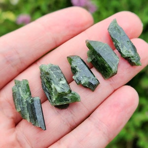 May include: A collection of six green, raw, and unpolished, crystals. The crystals are in various shapes and sizes, and they are all pointed at one end.