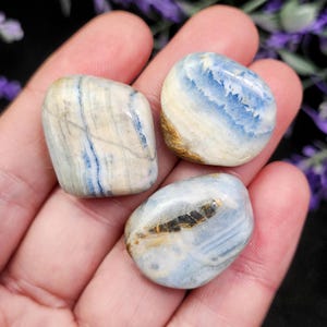May include: Three tumbled stones with a mix of blue, white, and tan colors. The stones are smooth and rounded, with visible patterns and variations in color. The stones are held in a hand.