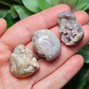 May include: Three agate stones with a variety of patterns and colors. The stones are a light brown, gray, and purple. The stones are smooth and have a natural, organic look.