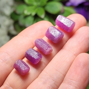 May include: Six pink, rough-cut, rectangular gemstones of varying sizes, arranged in a row on a hand.