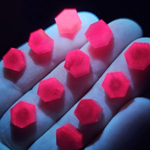 May include: A collection of 11 bright pink, faceted, hexagonal crystals. The crystals are arranged in a semi-circle on a hand.