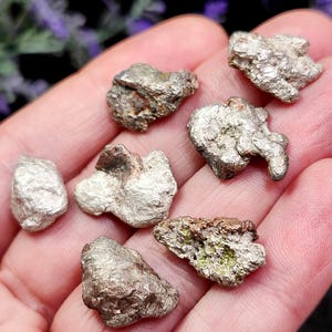 May include: A collection of irregularly shaped, silver-colored mineral specimens. The pieces vary in size and texture, with some showing a rough, crystalline surface and others appearing smoother. Some pieces have hints of brown and green.