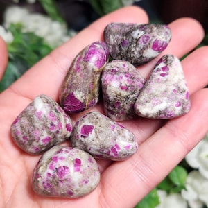 May include: A collection of seven polished tumbled stones featuring a mix of vibrant magenta-pink inclusions embedded within a smooth, mottled grey and white matrix. Each stone has a rounded, irregular shape with a glossy, reflective surface finish.