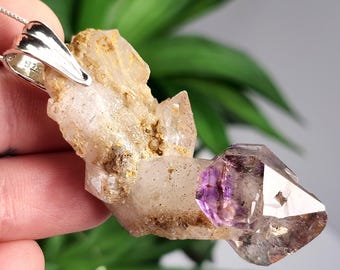 May include: A pendant featuring a raw, textured quartz crystal cluster with a prominent purple amethyst tip. The crystal is attached to a polished silver-toned metal bail stamped with 925, indicating sterling silver, and hangs from a thin chain.