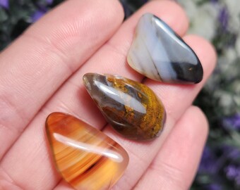 May include: Three agate stones with different patterns and colors. The first stone is a light gray and black agate with a swirling pattern. The second stone is a brown and yellow agate with a swirling pattern. The third stone is a red and orange agate with a swirling pattern.