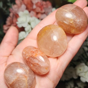May include: Four tumbled stones of clear quartz with orange and yellow inclusions. The stones are smooth and polished, and they are arranged in a row on a hand.