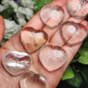 May include: Seven clear quartz heart-shaped crystals in various sizes, held in a hand. The crystals are translucent and have a slight shimmer.