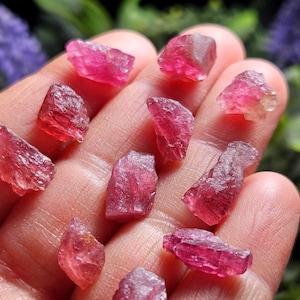 May include: A collection of small, raw, translucent pink tourmaline crystals with irregular, jagged edges and a vibrant magenta hue. These natural gemstone fragments exhibit a crystalline structure with varying levels of clarity and light reflection.