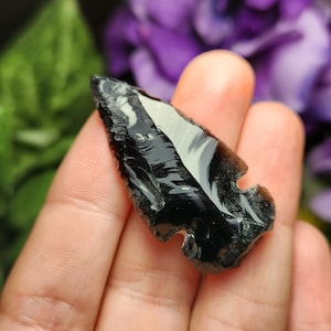 May include: A black obsidian arrowhead with white streaks, a type of volcanic glass, is held in a hand.