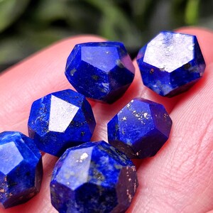 May include: A collection of six faceted lapis lazuli gemstones, each displaying a deep royal blue color with natural golden pyrite inclusions and dark mineral flecks, arranged in a cluster with geometric, multi-sided cut surfaces.