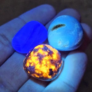 May include: Three glowing gemstones in a hand. One is a vibrant blue, another is a mix of orange and purple, and the third is a light blue with a dark streak. The stones are smooth and rounded, with a bright, almost neon, appearance.