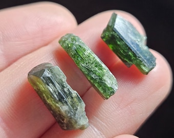 May include: Three green, faceted, crystal specimens. The crystals are long and narrow, with a slightly irregular shape. They are arranged in a row on a hand.
