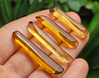 May include: Five amber-colored, elongated crystal points are displayed in a hand. The crystals have a smooth, polished appearance and vary in shades of yellow and brown. The crystals are arranged diagonally, with a blurred green background.