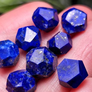 May include: A collection of small, faceted lapis lazuli gemstones with a deep blue color and visible gold-colored pyrite inclusions. Each stone features multiple geometric cut surfaces, creating a polished and reflective appearance.