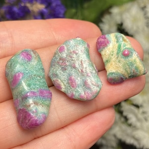 May include: Three raw ruby-in-zoisite stones. The stones display a mix of colours, including green, pink, and cream. Pink ruby crystals are visible throughout the green zoisite matrix. The stones are irregular in shape and appear natural.