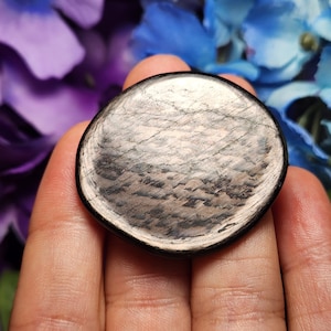 May include: A round, black obsidian stone with a silver sheen. The stone has a smooth, polished surface and a slightly irregular shape.