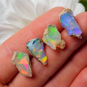 May include: Four rough, natural opal gemstones with iridescent colors, including green, blue, and pink, are displayed on a hand.