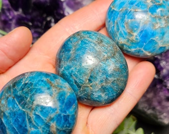 May include: Three blue apatite palm stones, each with a unique pattern of blue and white veins, are held in a hand. The stones are smooth and polished.