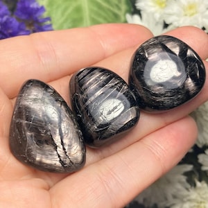 May include: Three black obsidian tumbled stones with silver streaks. The stones are smooth and polished.