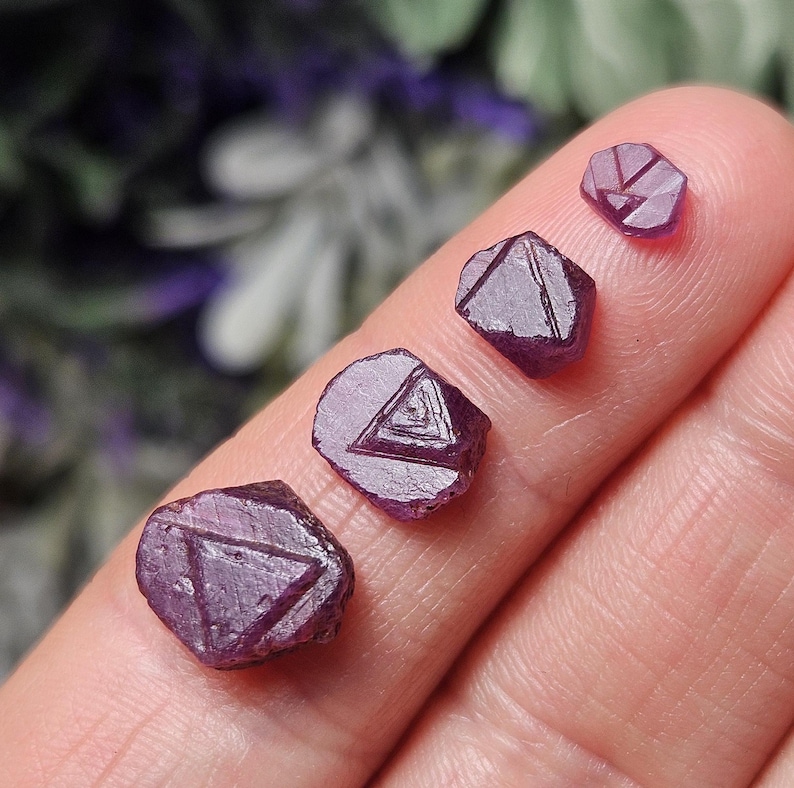 K&ouml;nnte beinhalten: Vier violette Rubinkristalle mit einem in jeden eingegrabenen dreieckigen Muster. Die Kristalle sind in einer Reihe vom gr&ouml;&szlig;ten zum kleinsten angeordnet.