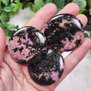 May include: Three polished, circular rhodonite stones featuring a mottled pattern of deep black and vibrant pink mineral inclusions. Each stone has a smooth, glossy surface that reflects light, showcasing the natural contrast between the dark and light colors.