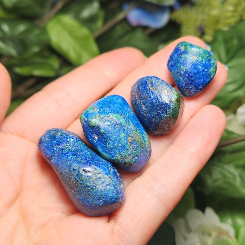 May include: Four polished azurite stones in varying shapes and sizes. The stones are a deep blue color with green and brown inclusions. The stones are held in an open hand, with a blurred green and white floral background.