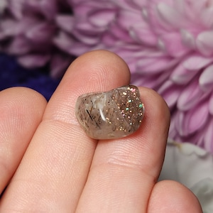 May include: A small, polished gemstone with a translucent base, speckled with dark inclusions and iridescent glitter. The stone is held in a hand, showcasing its natural, irregular shape. The gem's surface reflects light, creating a sparkling effect. The background is blurred, with hints of purple flowers.
