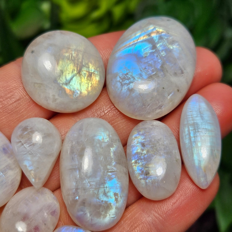 May include: A collection of iridescent moonstone cabochons in various shapes and sizes. The stones are a milky white color with a blue and green iridescent sheen.