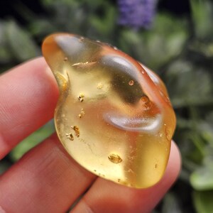 May include: A polished, translucent amber gemstone with a warm golden-yellow hue. The stone features natural internal inclusions, small golden flecks, and subtle reddish-brown streaks within its smooth, organic, and irregularly rounded shape.