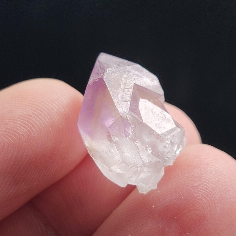 Puede incluir: Primer plano de un peque&ntilde;o cristal transparente con un tinte p&uacute;rpura en la parte superior. El cristal tiene m&uacute;ltiples facetas y se sostiene entre dos dedos sobre un fondo oscuro. Mineral natural.