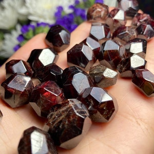May include: A collection of dark red garnet crystals, each with a faceted, geometric shape. The crystals are arranged in a pile on a hand.