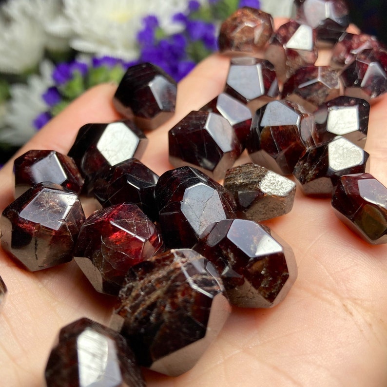 May include: A collection of dark red garnet crystals, each with a faceted, geometric shape. The crystals are arranged in a pile on a hand.
