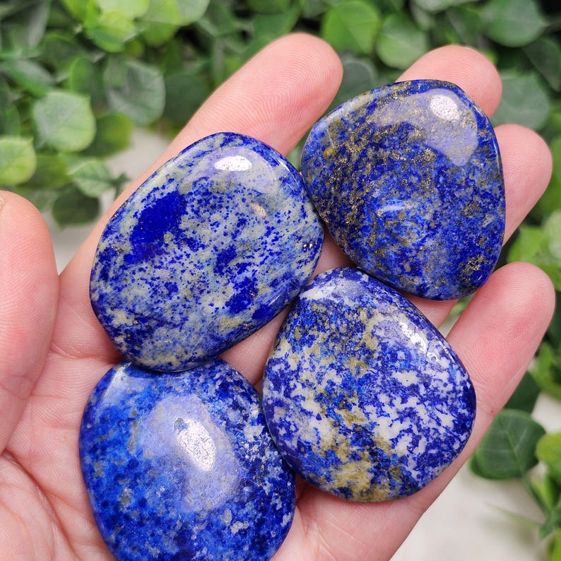 May include: Four polished lapis lazuli stones with a deep blue color and speckled with lighter blue and gold flecks. The stones are smooth and oval-shaped, perfect for jewelry making or collecting.