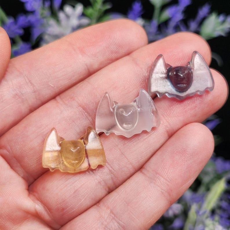 May include: Three small, translucent bat-shaped figurines in shades of yellow, clear, and purple are displayed. The bat figurines have detailed wings and faces. The background is blurred, with hints of purple flowers.