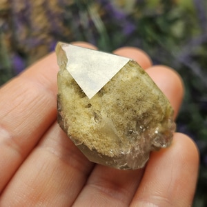 May include: A natural, raw crystal with a light brown and tan color. The crystal has a geometric shape with a clear, reflective surface on one side. The crystal is held in a hand, with a blurred background.