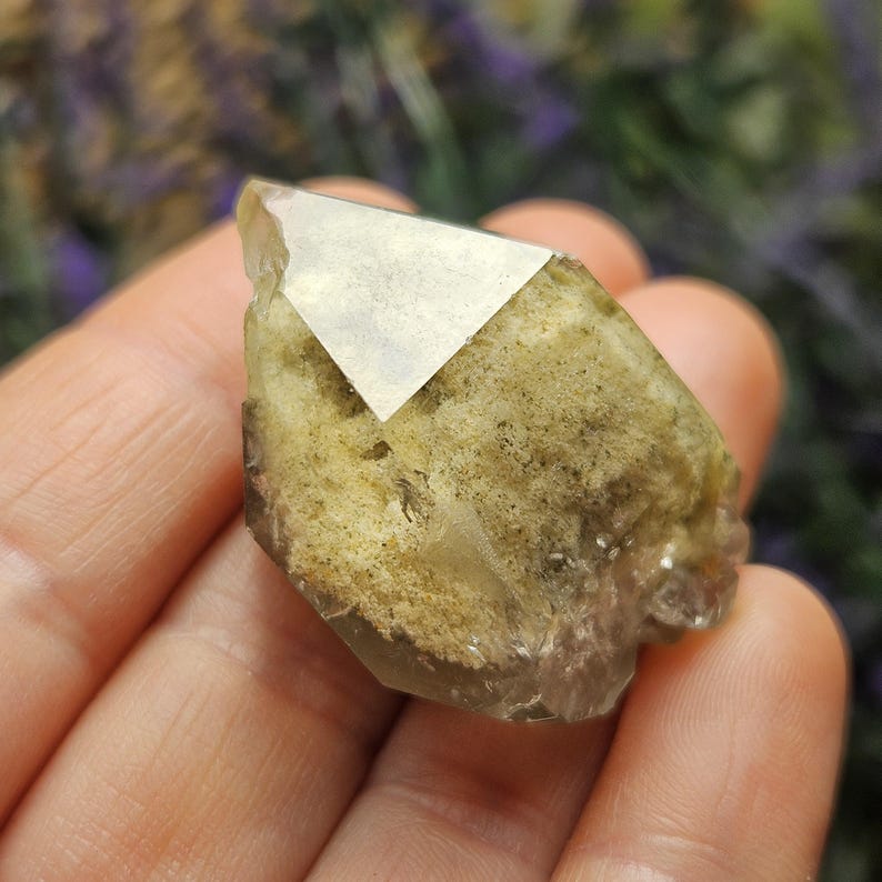 May include: A natural, raw crystal with a light brown and tan color. The crystal has a geometric shape with a clear, reflective surface on one side. The crystal is held in a hand, with a blurred background.