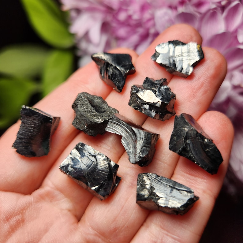 May include: A collection of eight black obsidian stones with a metallic sheen. The stones are various shapes and sizes, and some have a striated pattern.