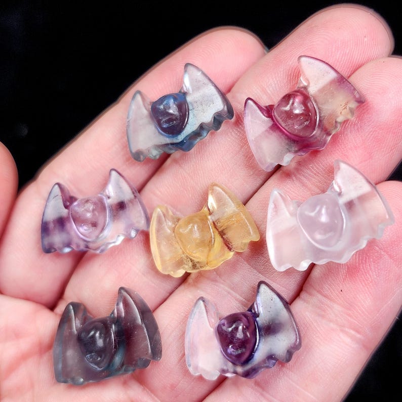 May include: A collection of small, carved gemstone bats in various colors, including purple, blue, yellow, and pink. The bats have detailed wing designs and a central body. The gemstones are translucent, allowing light to pass through, and are held in a hand.