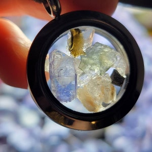 Puede incluir: Un colgante redondo con un marco negro, que muestra una colecci&oacute;n de gemas en bruto coloridas. Las piedras incluyen tonos de azul, verde y amarillo, creando una est&eacute;tica &uacute;nica y natural. El colgante cuelga de un cord&oacute;n negro.