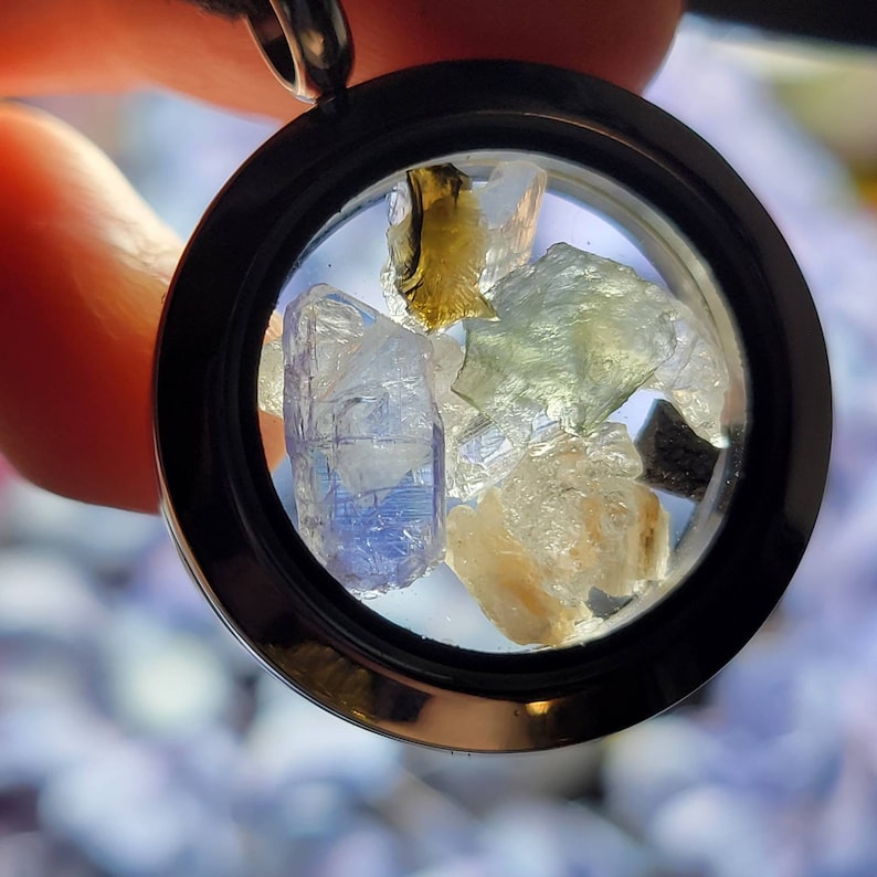 Puede incluir: Un colgante redondo con un marco negro, que muestra una colecci&oacute;n de gemas en bruto coloridas. Las piedras incluyen tonos de azul, verde y amarillo, creando una est&eacute;tica &uacute;nica y natural. El colgante cuelga de un cord&oacute;n negro.