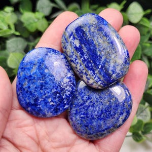 May include: Three polished lapis lazuli stones with a deep blue color and gold flecks. The stones are smooth and oval-shaped, with a glossy finish. The stones are held in a hand.