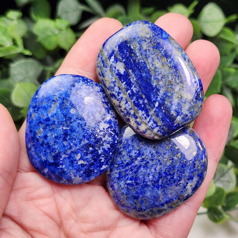 May include: Three polished lapis lazuli stones with a deep blue color and gold flecks. The stones are smooth and oval-shaped, with a glossy finish. The stones are held in a hand.