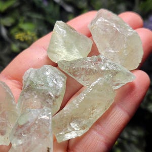 May include: A collection of pale green, translucent rough-cut crystals. The crystals have irregular shapes and varying sizes, with a slightly frosted appearance. The crystals are held in a hand, with a blurred green background.