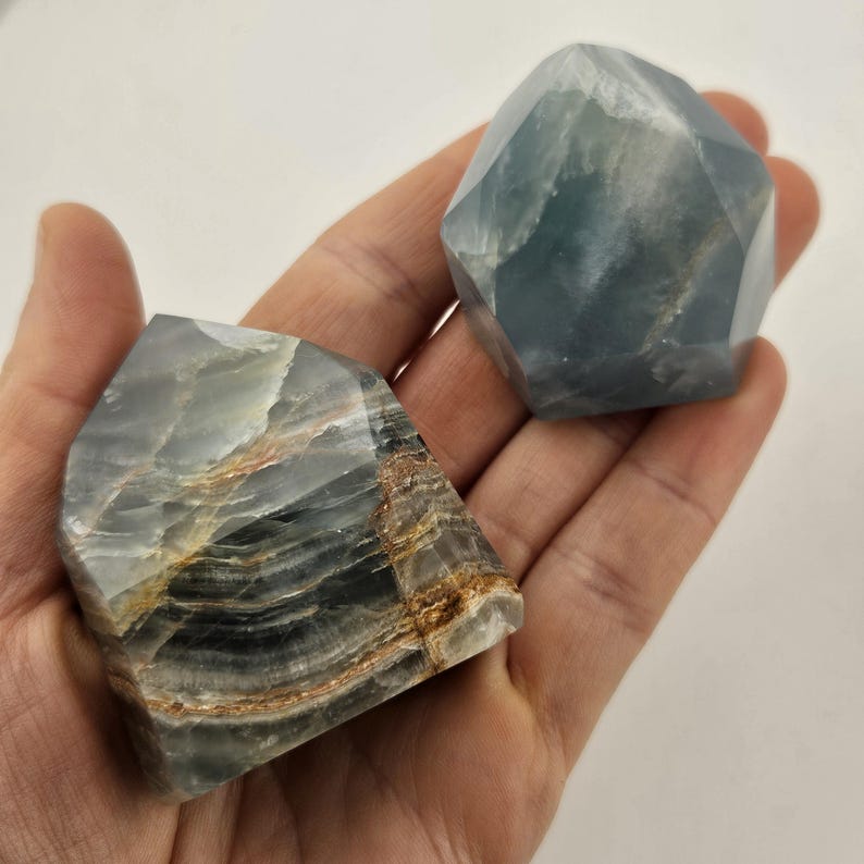 May include: Two polished, faceted gemstone sculptures. One is a light blue-green colour, and the other is a mix of grey, brown, and tan. The stones have a smooth, reflective surface and are held in a hand.