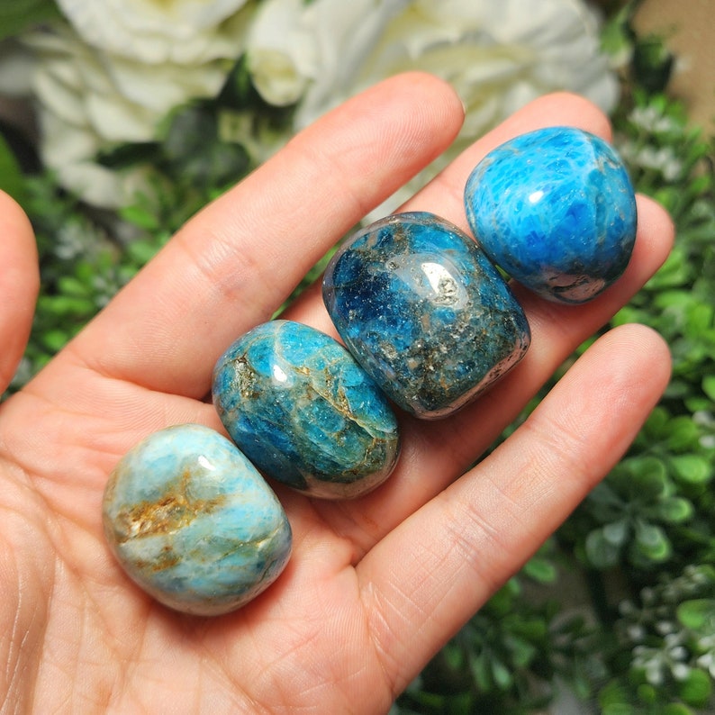 May include: Four polished, oval-shaped gemstones in shades of blue and teal are held in a hand. The stones have a smooth, glossy texture with variations in color and some with gold-colored inclusions. The background is blurred with greenery.