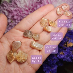 May include: A hand holds several polished, golden-brown gemstones. Text labels indicate sizes: X-Small (.3g-.5g), Small (.5g-1g), Medium (1g-2g), and Large (2g-4g). The gemstones are translucent with a sparkling effect, varying in size and shape.