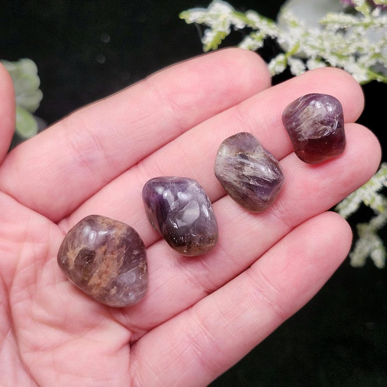 May include: Four polished amethyst stones in varying shades of purple and brown are displayed in a hand. The stones have a smooth, rounded shape and show natural patterns. The background is dark, highlighting the stones.