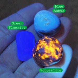May include: Three glowing gemstones: Green Fluorite, Blue Amber, and Yooperlite. The Green Fluorite is a vibrant blue, the Blue Amber is a lighter blue, and the Yooperlite has a dark base with bright orange and yellow inclusions. The stones are held in a hand.