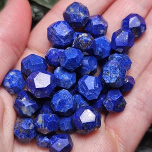 May include: A collection of small, faceted lapis lazuli gemstones. Each stone features a deep royal blue color with natural golden pyrite inclusions and a multi-faceted geometric cut that creates sharp edges and reflective surfaces across the group.